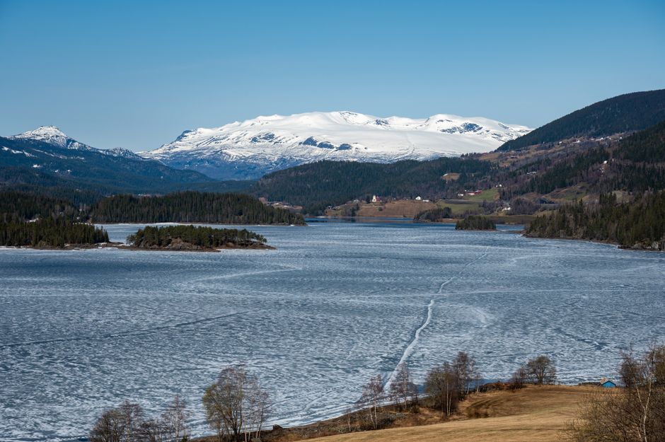 Scenic winter view of frozen lake and snow-capped mountains in Aurdal, Norway, under clear blue sky.