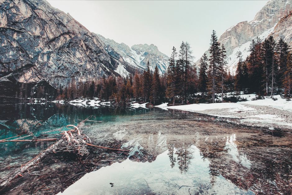 A tranquil winter scene of a clear lake surrounded by snowy mountains and pine trees.